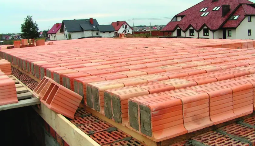 Budowa stropu ackermana. Czerwone pustaki ceramiczne ułożone w rzędach, tworzące konstrukcję stropu. W tle domy z czerwonymi dachami.
