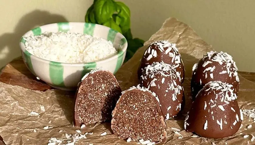 Hemmagjorda delicatobollar, täckta med choklad och kokosflingor, redo att avnjutas. En skål med kokosflingor står bredvid.