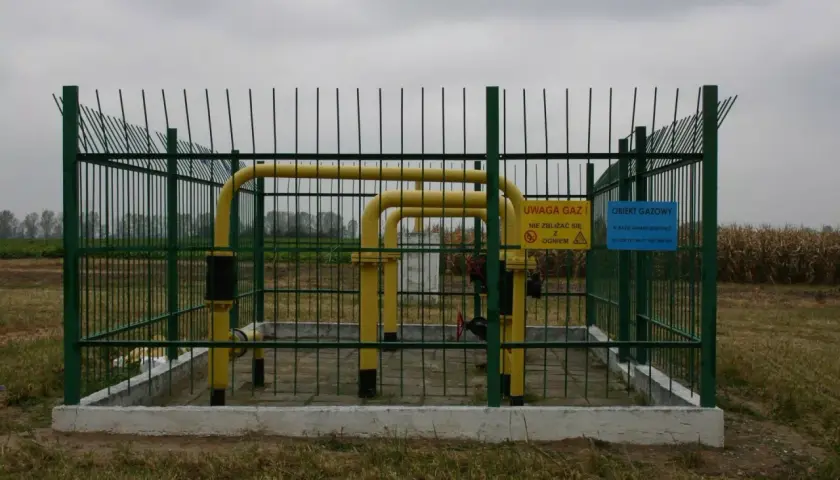 Ogrodzony obiekt gazowy z żółtymi rurami. Tablice ostrzegawcze informują o zagrożeniu i numerach alarmowych. Widać, że odległość budynku od gazociągu średniego ciśnienia jest bezpieczna.