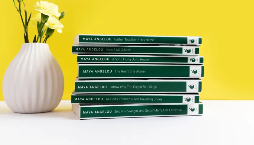 Stos **książek** **Maya Angelou** obok wazonu z żółtymi kwiatami.