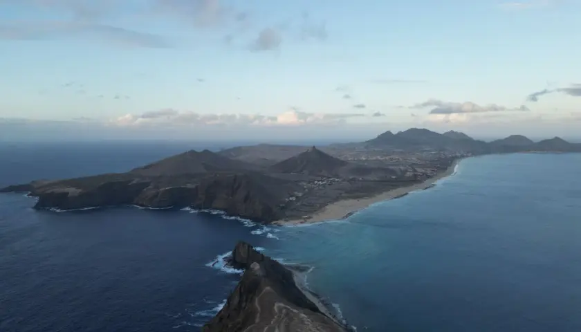 Dramatic coastline of Porto Santo island, with rugged cliffs meeting turquoise waters and a long sandy beach stretching towards distant mountains under a cloudy sky.