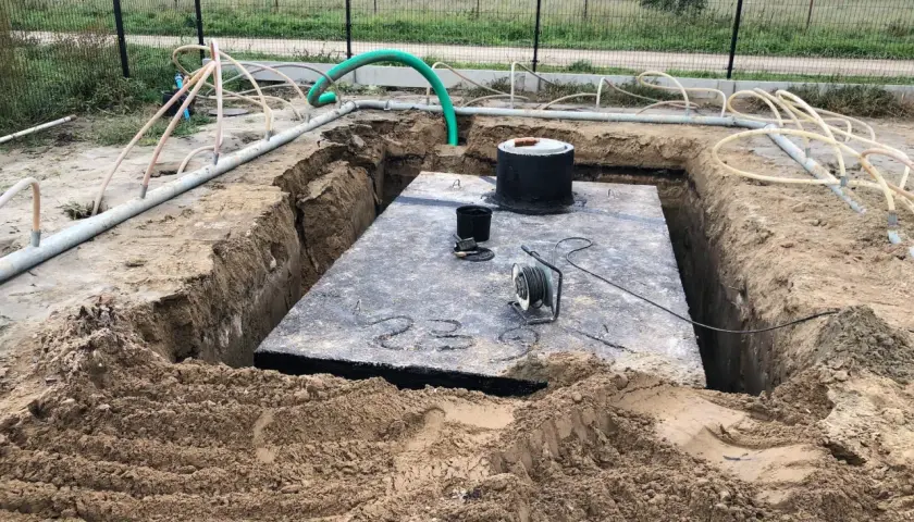 Widok na wykop pod szambo. Duża betonowa płyta, okrągły właz i liczne rury.