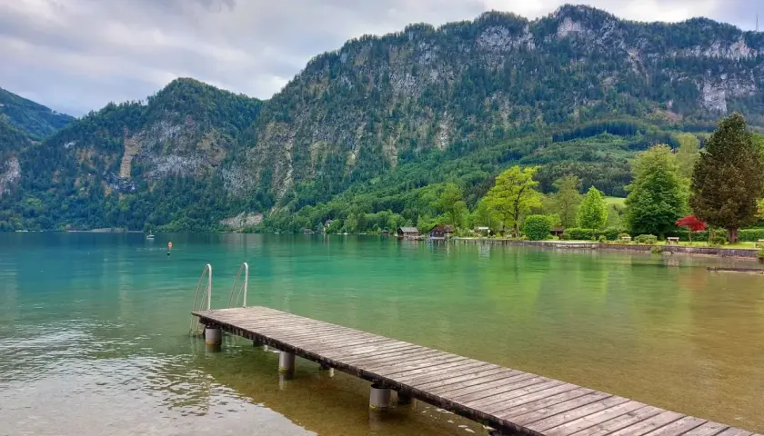 Idealne miejsce na wakacje w **Austrii latem z dziećmi**. Drewniane molo prowadzi do krystalicznie czystej wody, a w tle wznoszą się zielone góry.
