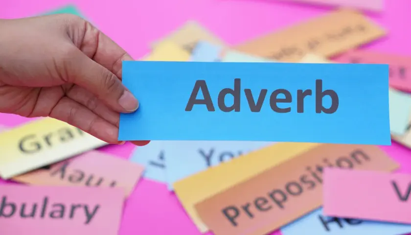 Una mano tiene un biglietto blu con la parola "Adverb". Sullo sfondo, altri biglietti colorati con parole come "Grammar" e "Vocabulary".