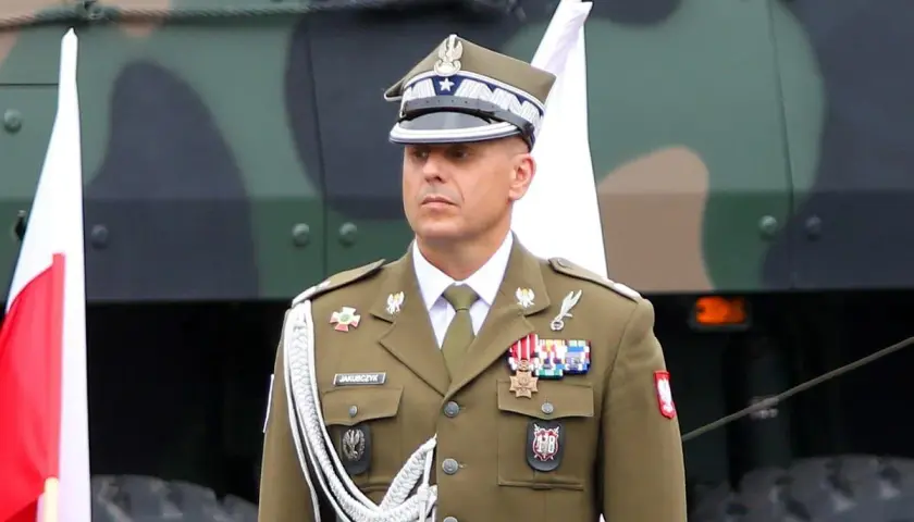 Oficer w mundurze, z orderami na piersi, stoi obok polskiej flagi. Nad nim widać fragment pojazdu wojskowego.