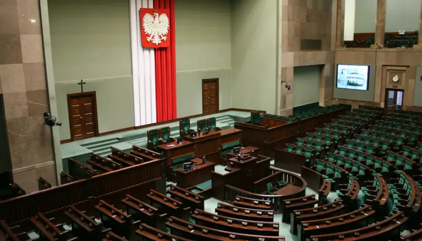 Sala plenarna Sejmu RP z godłem Polski i rzędami zielonych foteli.