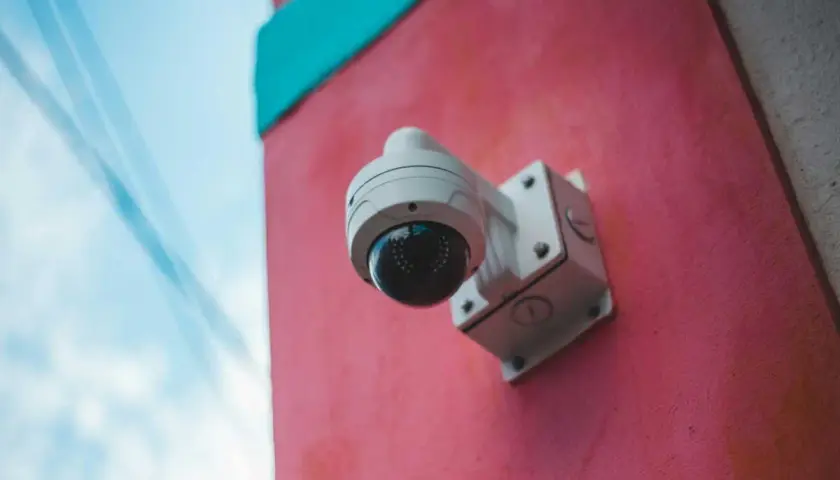 Caméra de surveillance blanche fixée sur un mur rose vif, sous un ciel bleu. Un rappel visuel de la loi sur la caméra de surveillance.