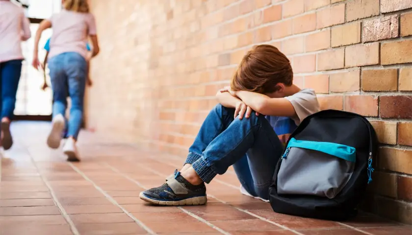 Chłopiec siedzi przy ceglanej ścianie, ukrywając twarz w dłoniach. W tle oddalają się inne dzieci. To obraz bullying po polsku.