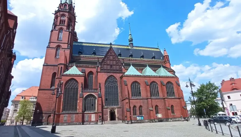 Gotycka katedra w mieście nad Kaczawą, z ceglaną fasadą i zielonymi dachami, na tle błękitnego nieba z chmurami.