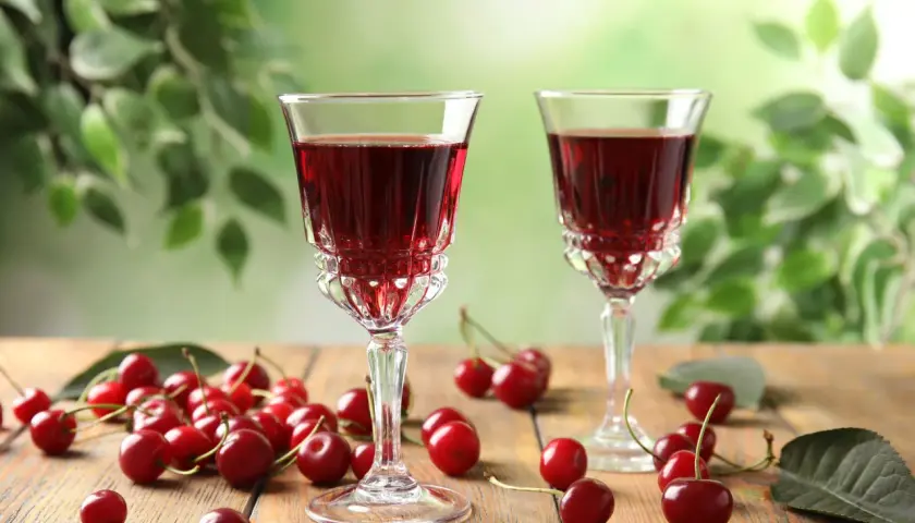 Dwa kieliszki z rubinowym alkoholem z wiśni, otoczone świeżymi owocami i zielonymi liśćmi.