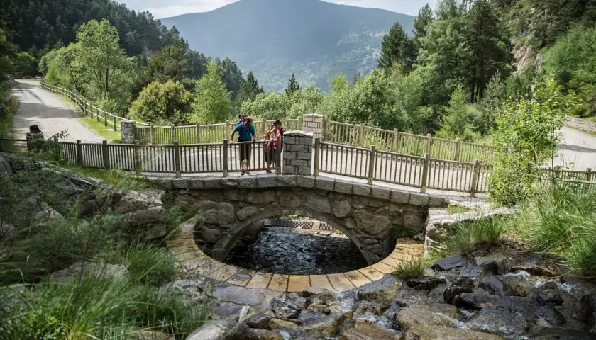 Pareja en un puente de piedra sobre un arroyo en Les Pardines, Andorra. Montañas y bosques al fondo.