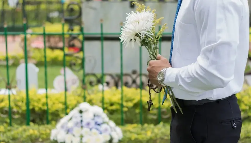 Mężczyzna w białej koszuli i niebieskim krawacie trzyma bukiet białych kwiatów i różaniec. W tle cmentarz, gdzie spoczywaj w pokoju.