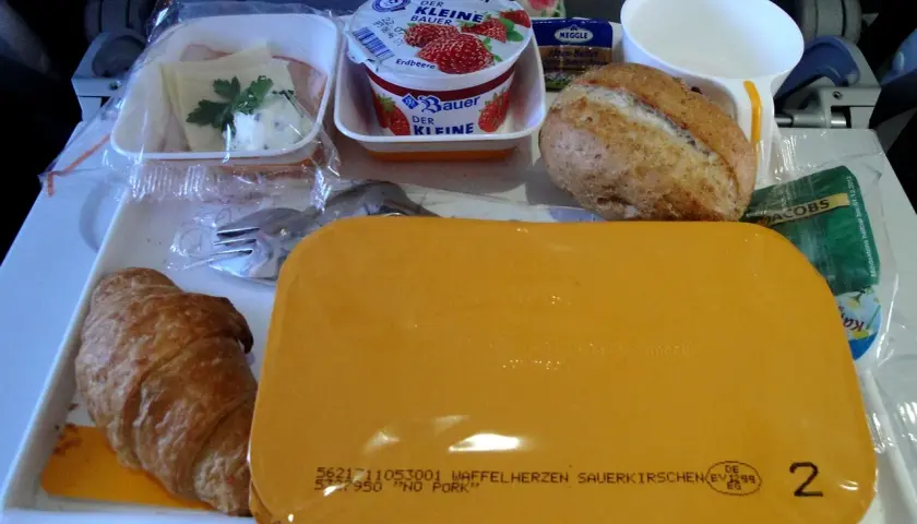 Taca z jedzeniem w samolocie: rogalik, jogurt, bułka, masło, kawa i żółty pojemnik.