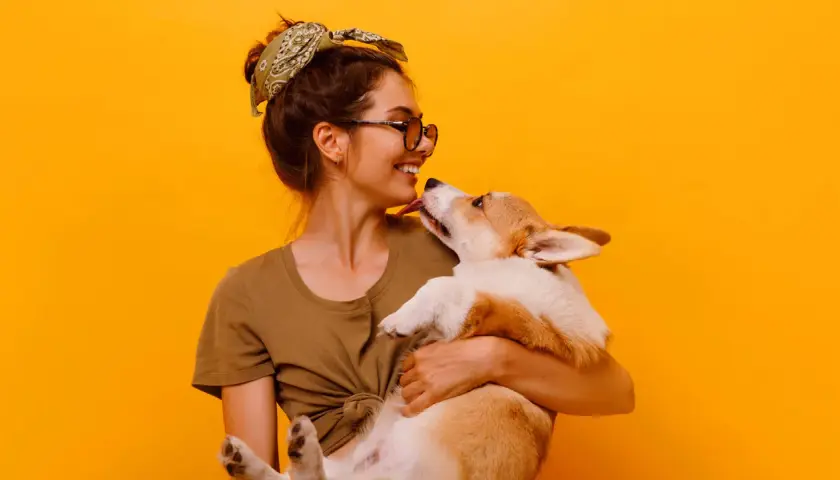 Uśmiechnięta dziewczyna z okularami i bandaną na głowie tuli corgi. Piesek liże ją po nosie. Wiele imion na N, jak Nela czy Nika, pasowałoby do tej słodkiej pary.