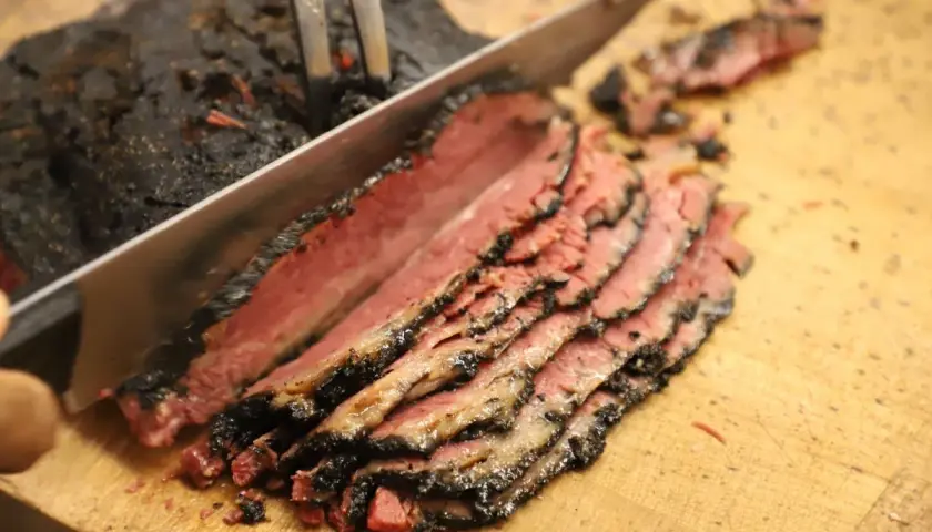 Un cuchillo corta un trozo de pastrami ahumado y jugoso, revelando su interior rosado.