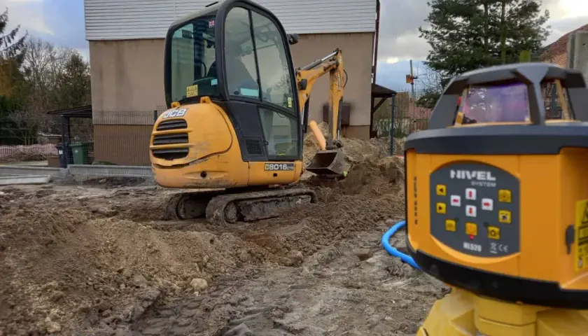Żółty koparko-ładowarka i niwelator laserowy na placu budowy. To narzędzia, które pomagają w precyzyjnym wyznaczaniu poziomów terenu.