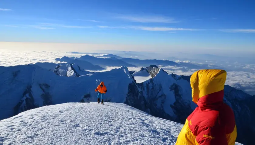 Widok z góry, gdzie są Himalaje. Dwóch alpinistów w kolorowych kurtkach na ośnieżonym szczycie, pod nimi morze chmur i kolejne pasma górskie.