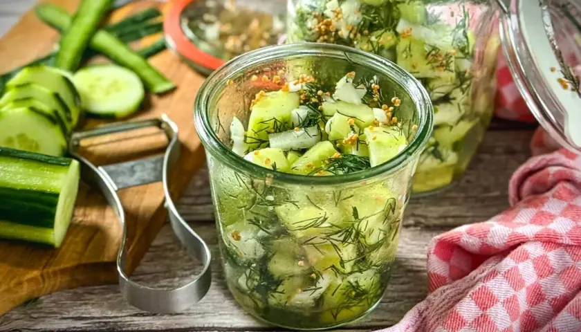 Frischer gurkensalat ohne zucker, zubereitet mit Dill und Senfkörnern, in Gläsern abgefüllt. Gurken und Sparschäler liegen bereit.