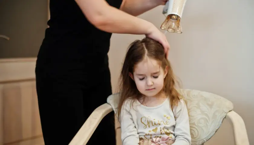 Mama suszy włosy córki suszarką, dbając o delikatną pielęgnację włosów dziecka.