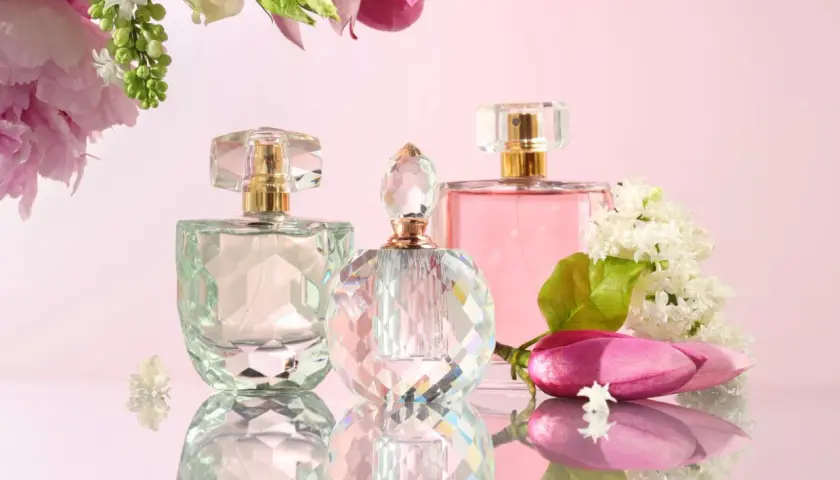 Trzy eleganckie flakoniki perfum na różowym tle, ozdobione kwiatami.