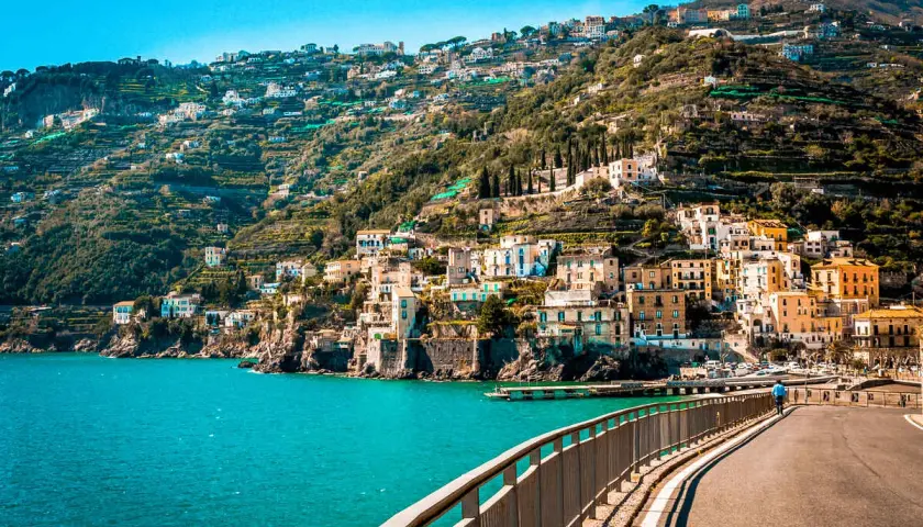 A scenic road curves along the coast, offering breathtaking views of colorful buildings clinging to a hillside. The turquoise sea sparkles below, a perfect backdrop for an unforgettable Amalfi drive.
