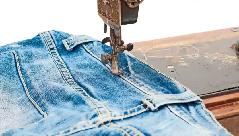 A sewing machine stitches a classic jean stitch onto blue denim fabric, highlighting the durable construction of jeans.
