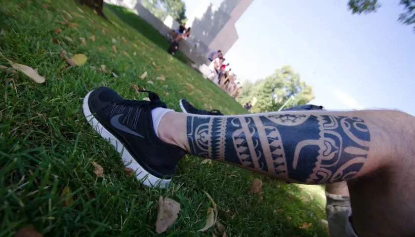 Noga z imponującym tatuażem polinezyjskim, w czarnych butach sportowych, na tle zielonej trawy i ludzi w oddali.