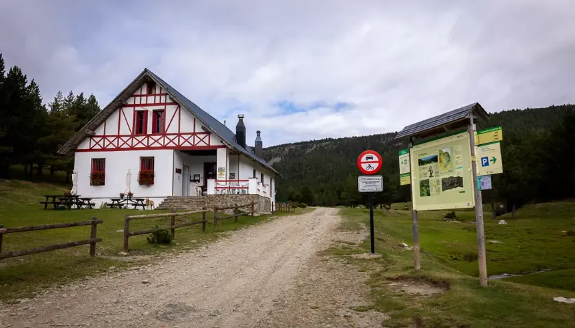 Refugi comes de rubio, un refugio de montaña blanco con detalles rojos, rodeado de pinos y un camino de grava.