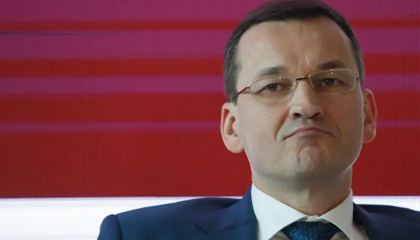 Mateusz Morawiecki w garniturze i okularach na tle czerwono-fioletowych pasów.