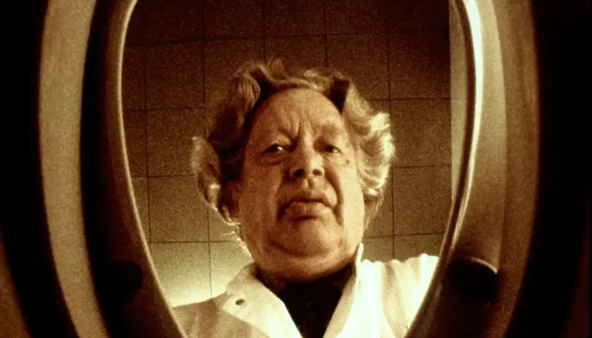 En man med lockigt hår, klädd i vit rock, tittar in i en toalettstol. Ernst Hugo Järegård i en minnesvärd scen.
