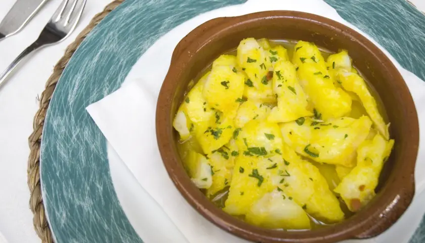 Delicioso bacalao a la cazuela, con trozos tiernos de pescado en una salsa dorada y perejil picado, servido en una cazuela de barro.