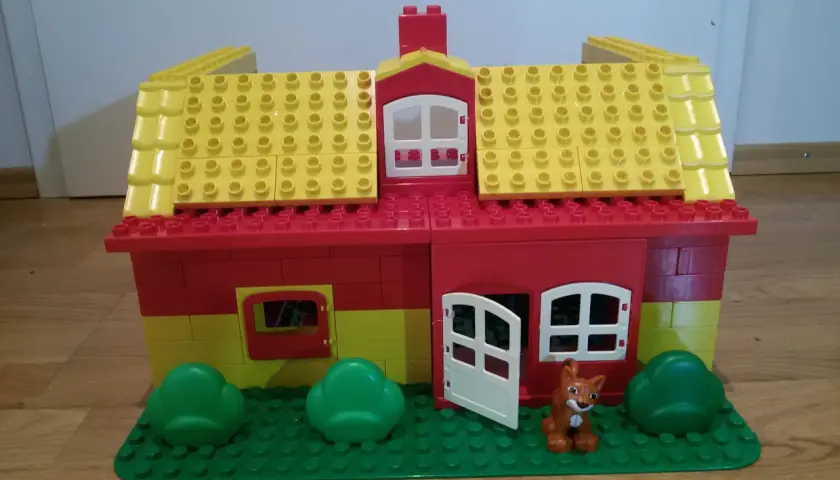Ein farbenfrohes Lego Duplo Haus mit gelbem Dach, roten Wänden, Fenstern, einer Tür und einem kleinen Fuchs davor. Perfekt für kreative Bauanleitungen.