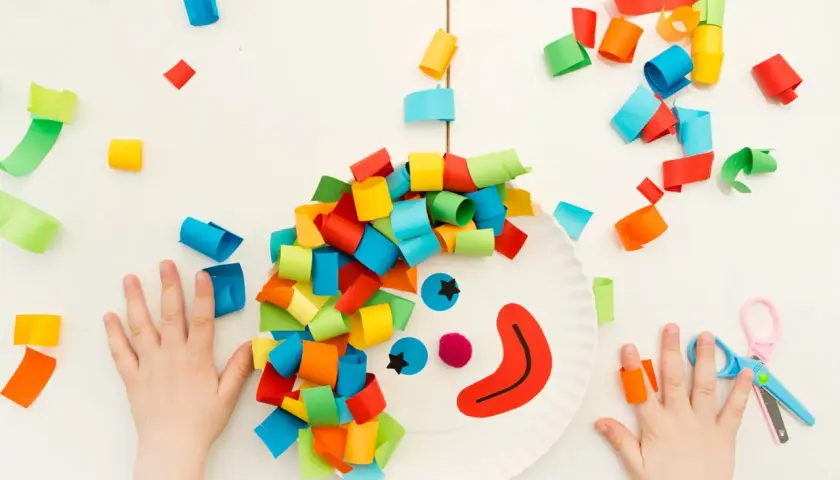 Bambini creano lavoretti di carnevale da appendere: un pagliaccio con capelli colorati su un piatto di carta, circondato da coriandoli di carta.