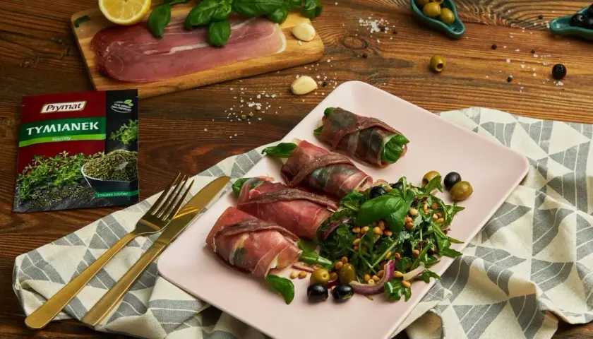 Roladki z szynką parmeńską i anchois, podane z sałatką z rukoli, oliwek i orzeszków pinii. Obok leży cytryna, czosnek i paczka tymianku.