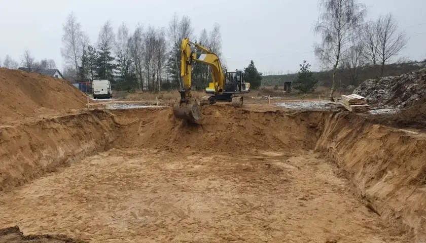 Żółta koparka pracuje przy wykopy pod fundamenty. W tle drzewa i biały van.