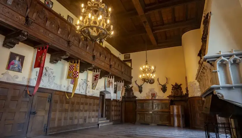 Wnętrza Zamku Czocha: bogato zdobiona sala z drewnianymi panelami, galerią, żyrandolami i trofeami łowieckimi.