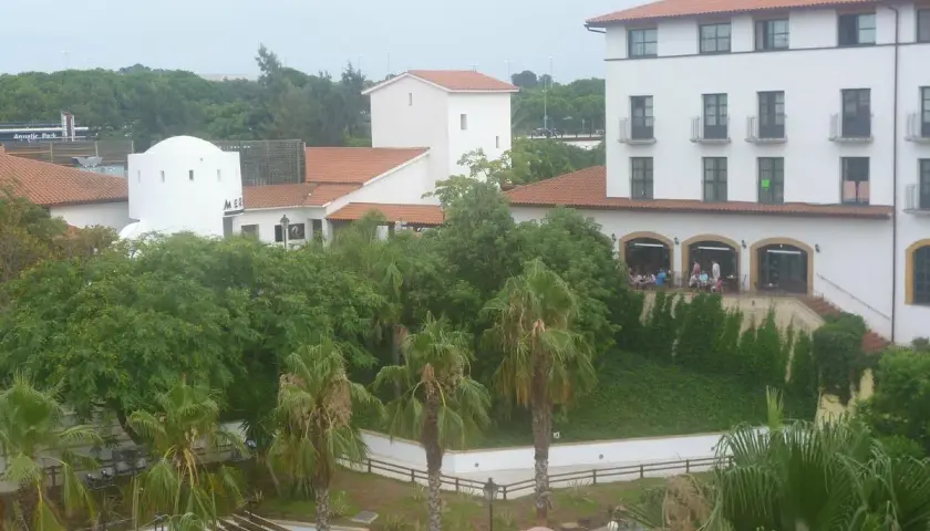 Edificio blanco con tejado naranja, palmeras y vegetación exuberante. Parece el hotel El Paso PortAventura, listo para recibir a sus huéspedes.