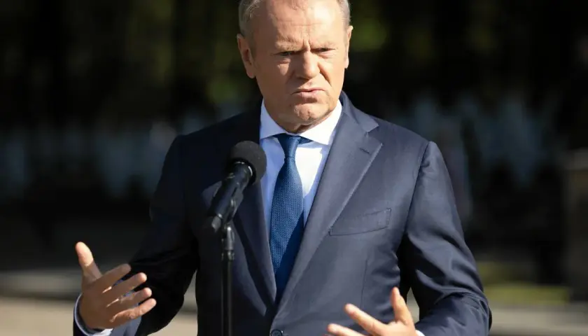 Donald Tusk w garniturze przemawia przy mikrofonie, gestykulując rękami.
