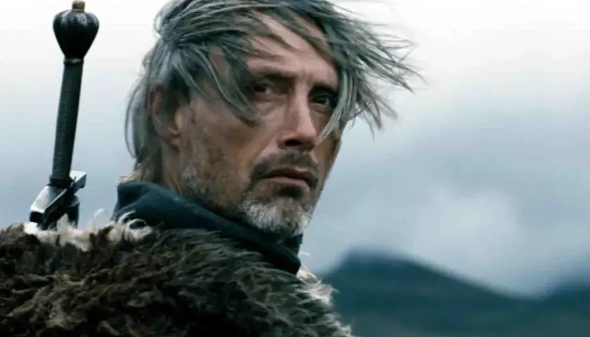 Mads Mikkelsen młody, z mieczem za plecami i w futrzanym płaszczu, spogląda w dal.
