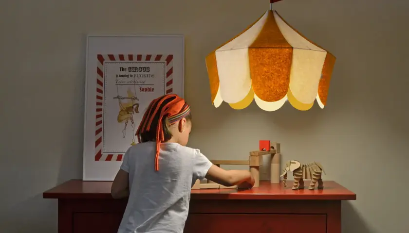Una niña juega con bloques de madera bajo una lámpara colgante infantil con forma de carpa de circo.
