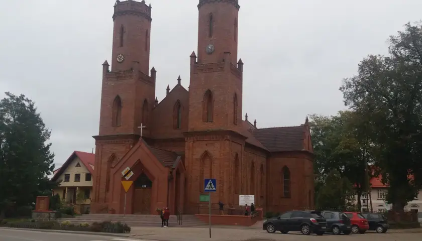 Imponujący kościół w Krokowej z cegły, z dwiema wieżami i zegarami, otoczony zielenią i samochodami.
