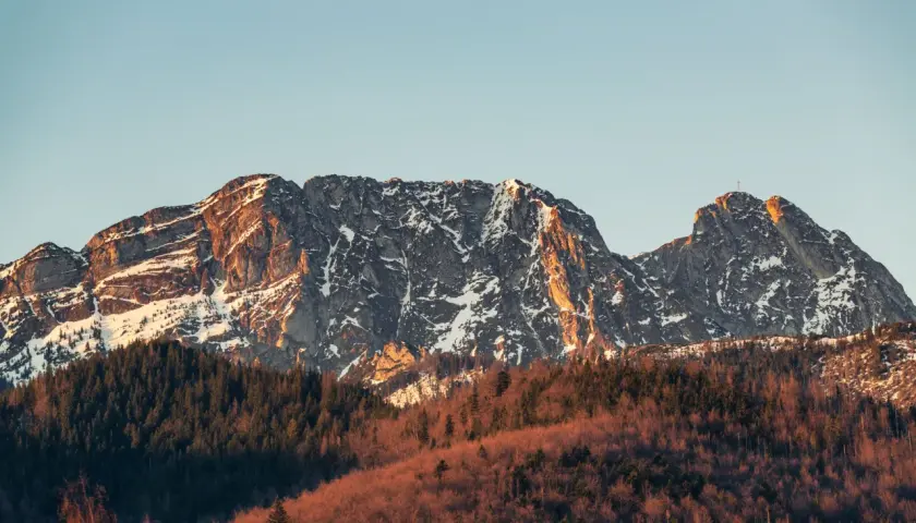 Giewont w słońcu, z ośnieżonymi zboczami i lasem u podnóża. Jego imponująca wysokość robi wrażenie.