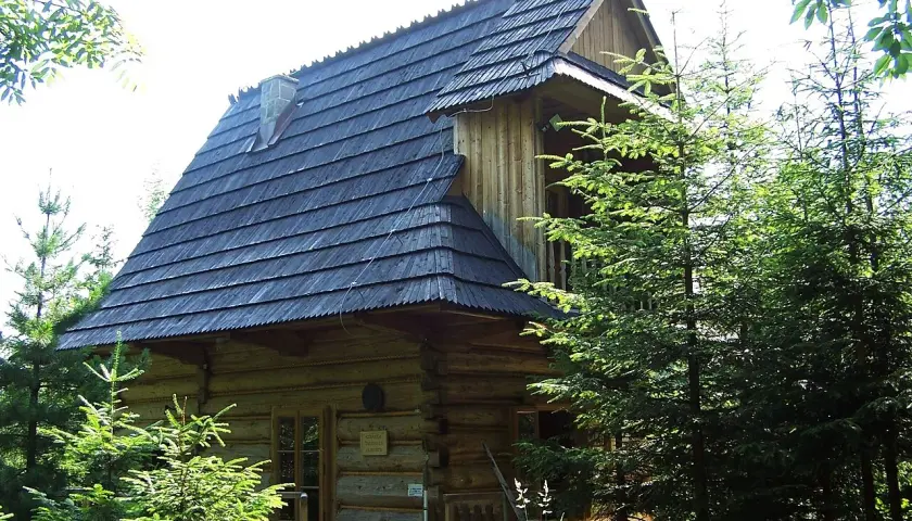 Drewniana pustelnia Brata Alberta, otoczona zielenią, z charakterystycznym gontowym dachem i kamiennym kominem.