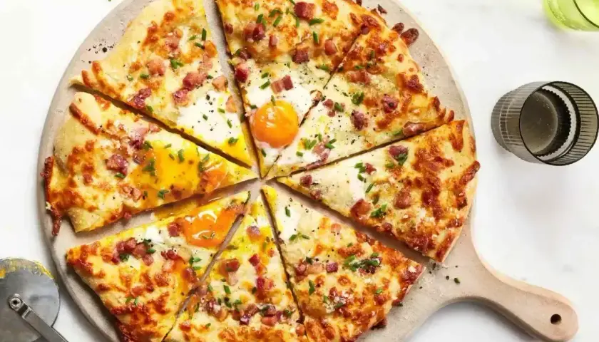 Kawałki pizzy carbonara z jajkiem sadzonym, boczkiem i szczypiorkiem na desce.