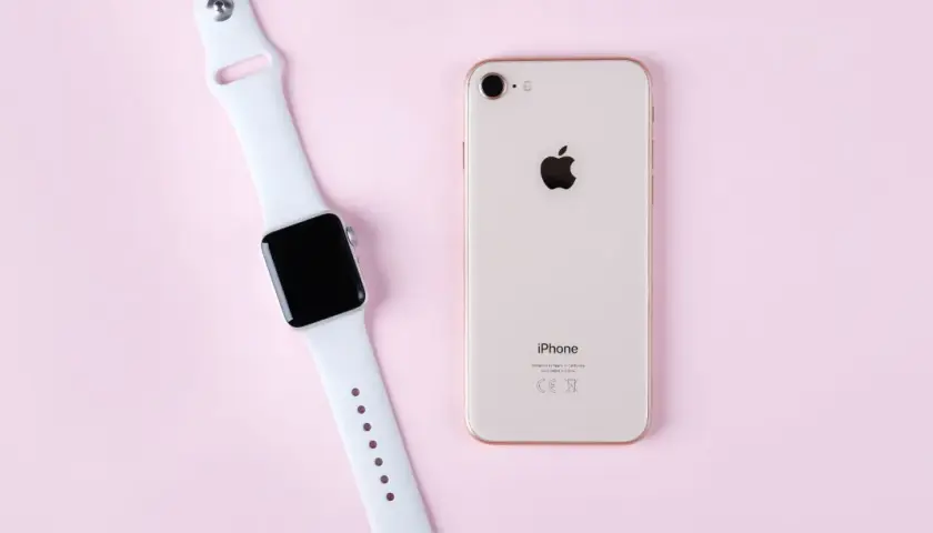 Apple Watch und iPhone liegen nebeneinander auf rosa Untergrund, bereit zum Laden.
