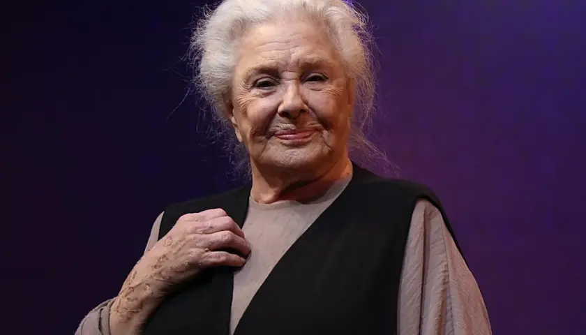 Lola Herrera, con su melena plateada y una sonrisa serena, luce un chaleco negro sobre una blusa beige. Su mano izquierda, con un anillo, reposa sobre su pecho.