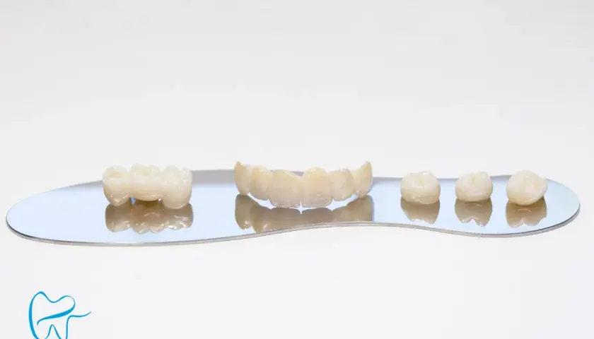 Zęby ceramiczne, w tym mostek i pojedyncze korony, prezentowane na lustrzanej powierzchni. Wykonane z materiałów takich jak korona cyrkonowa, zapewniają estetykę i trwałość.