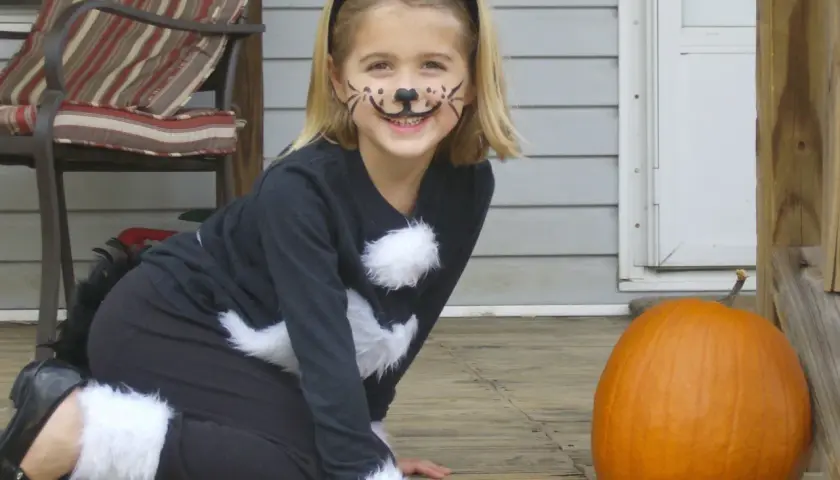 Bambina con trucco da gatto e orecchie, pronta per vestiti carnevale veloci fai da te bambini. Zucca arancione accanto.