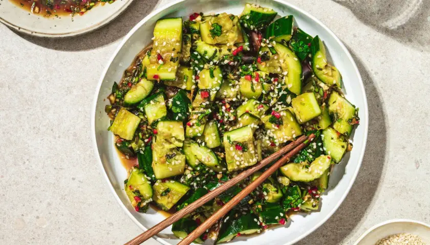 En skål med krossad gurksallad, toppad med sesamfrön och färska örter, redo att avnjutas med ätpinnar.