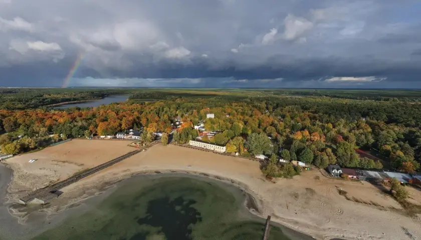 Jesioro Skorzęcin jesienią. Tęczowy łuk nad ciemnymi chmurami, piaszczysta plaża i kolorowy las.